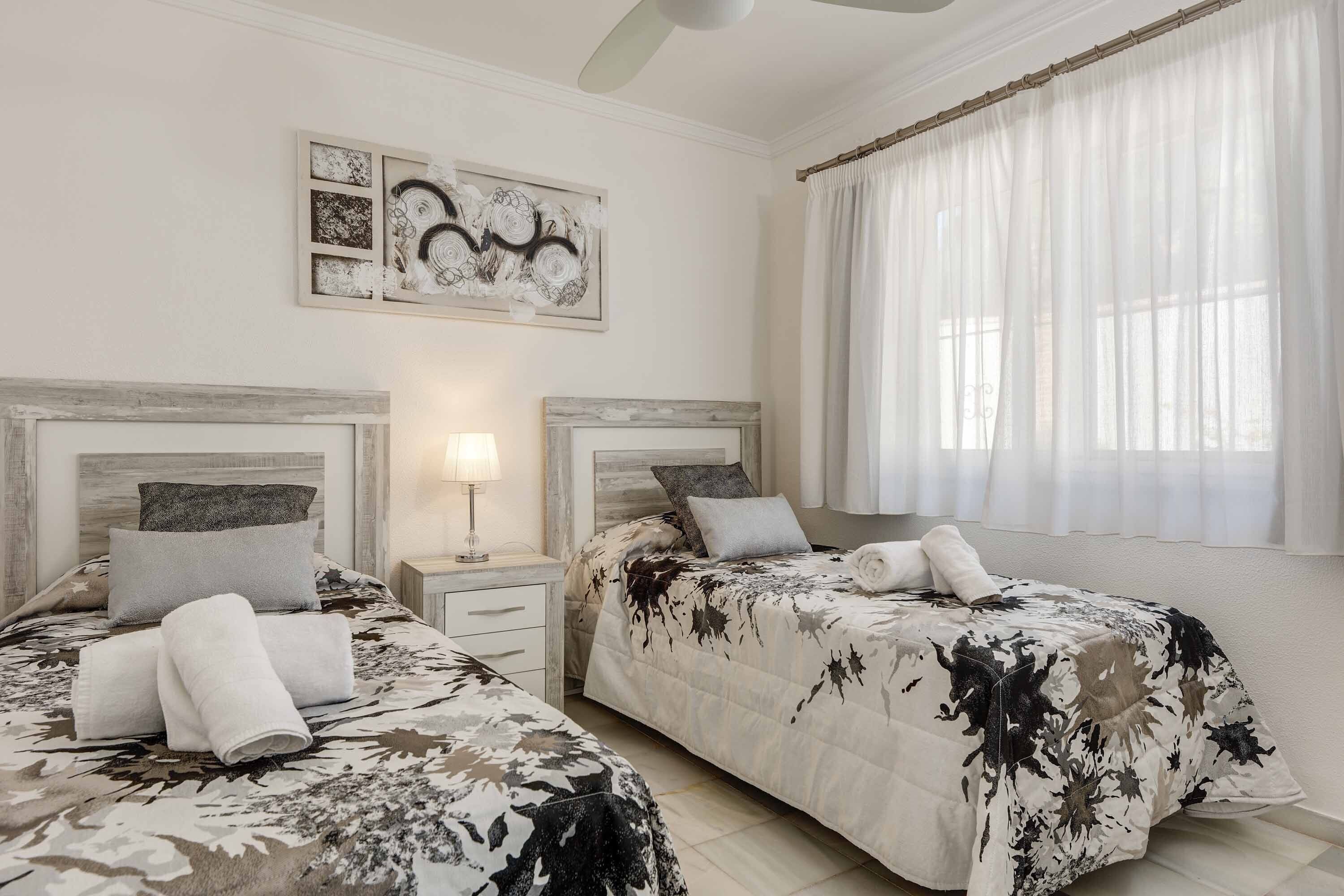 A hotel room with two beds, a nightstand, a lamp, and a large window with sheer curtains.