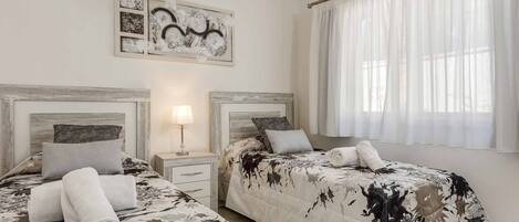 A hotel room with two beds, a nightstand, a lamp, and a large window with sheer curtains.
