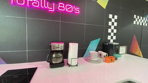Una estación de café moderna con un letrero de neón que dice “Totally 80's”, una cafetera, un dispensador de servilletas de papel y varias tazas y vasos sobre una encimera blanca.