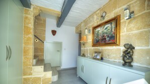 Un pasillo con pared de piedra, una escalera, una puerta blanca, un armario con lámpara y un cuadro en la pared.
