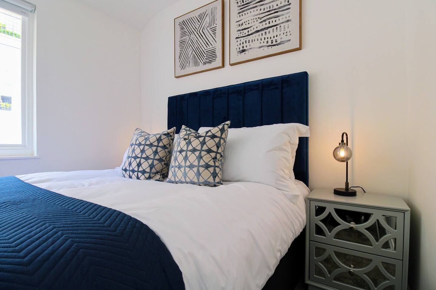 A neatly made bed with a blue headboard, white bedding, and patterned pillows. There is a bedside table with a lamp and a decorative wall piece above it.
