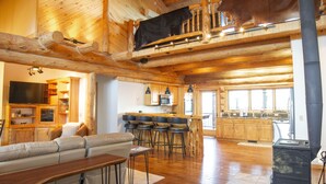 Amplia sala de estar con techo alto, vigas de madera, sofá, mesa de comedor y cocina con armarios de madera y estufa.