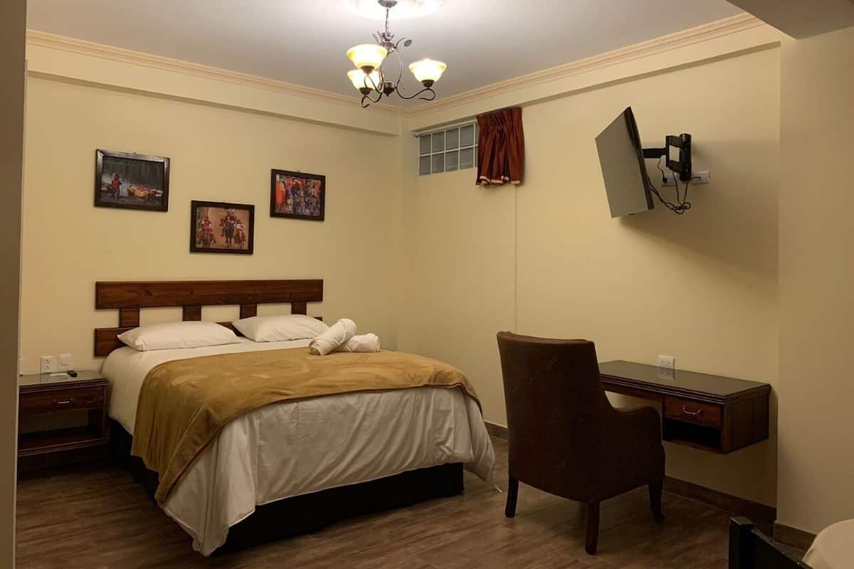 A bedroom with a bed, a desk, a chair, a TV mounted on the wall, and a ceiling light fixture.