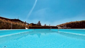 Ein klarer, blauer Pool mit einer Fontäne in der Mitte, umgeben von einer Hecke und einem Gebäude mit einem geneigten Dach.