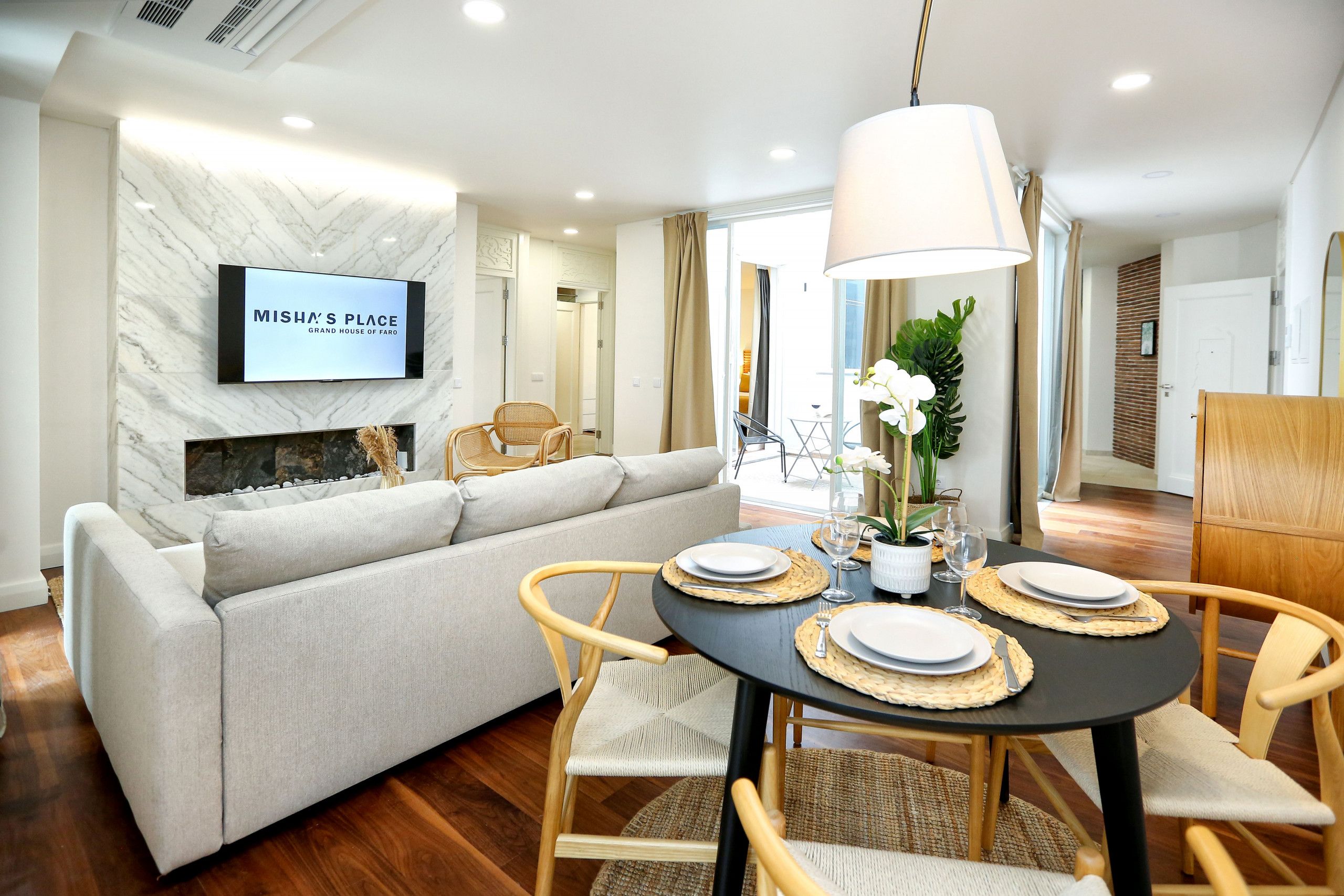 A modern living room with a sofa, a dining table set for two, and a flat-screen TV mounted on a marble wall.