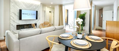 A modern living room with a sofa, a dining table set for two, and a flat-screen TV mounted on a marble wall.