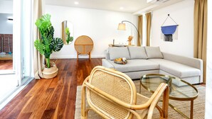 Ein modernes Wohnzimmer mit einer Couch, einem Glastisch, einem Rattanstuhl, einer Topfpflanze und einem blauen Wandbehang.