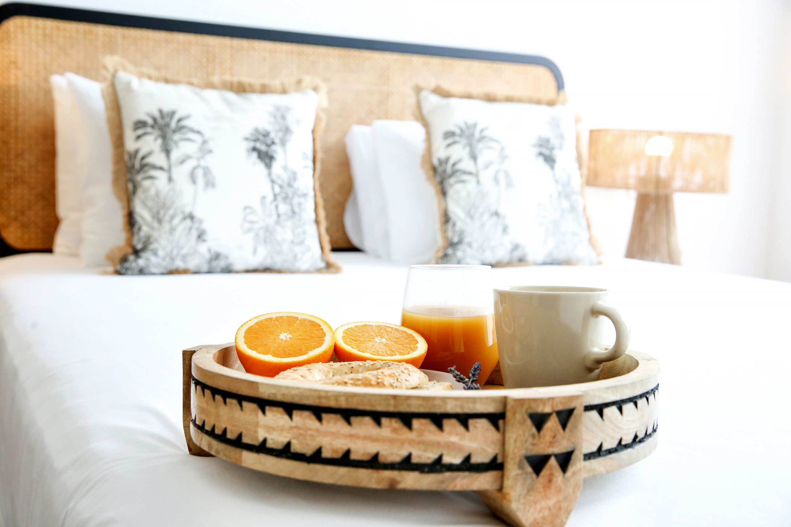 A breakfast tray with orange slices, a glass of orange juice, and a cup on a bed with white bedding and patterned pillows.
