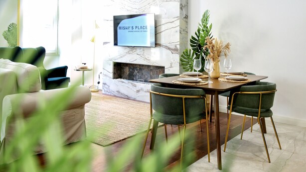 A modern living room with a fireplace, a dining area with green chairs, and a TV displaying "Misha's Place". - GREEN FARO : MODERN DUPLEX WITH A BLISS TERRACE (Faro)