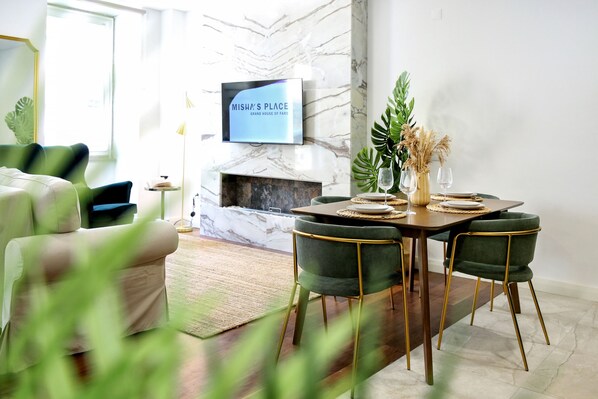 A modern living room with a fireplace, a dining area with green chairs, and a TV displaying "Misha's Place".