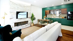 A modern living room with a white sofa, a dining area with a table and chairs, a kitchen with green cabinets, and a mounted TV displaying "Misha's Place Grand House of Trees". - GREEN FARO : MODERN DUPLEX WITH A BLISS TERRACE (Faro)