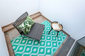 A corner of a room with a patterned floor mat, a chair with a cushion, a small table with a book and a cup.