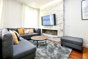 A modern living room with a grey sofa, a round coffee table, and a grey ottoman. A flat-screen TV is mounted on a marble wall above a fireplace.