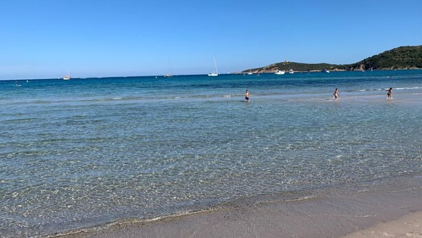 A beach with clear water, boats in the distance, and a hilly landscape. - Rêves de mer en Tout Compris (Linguizzetta)