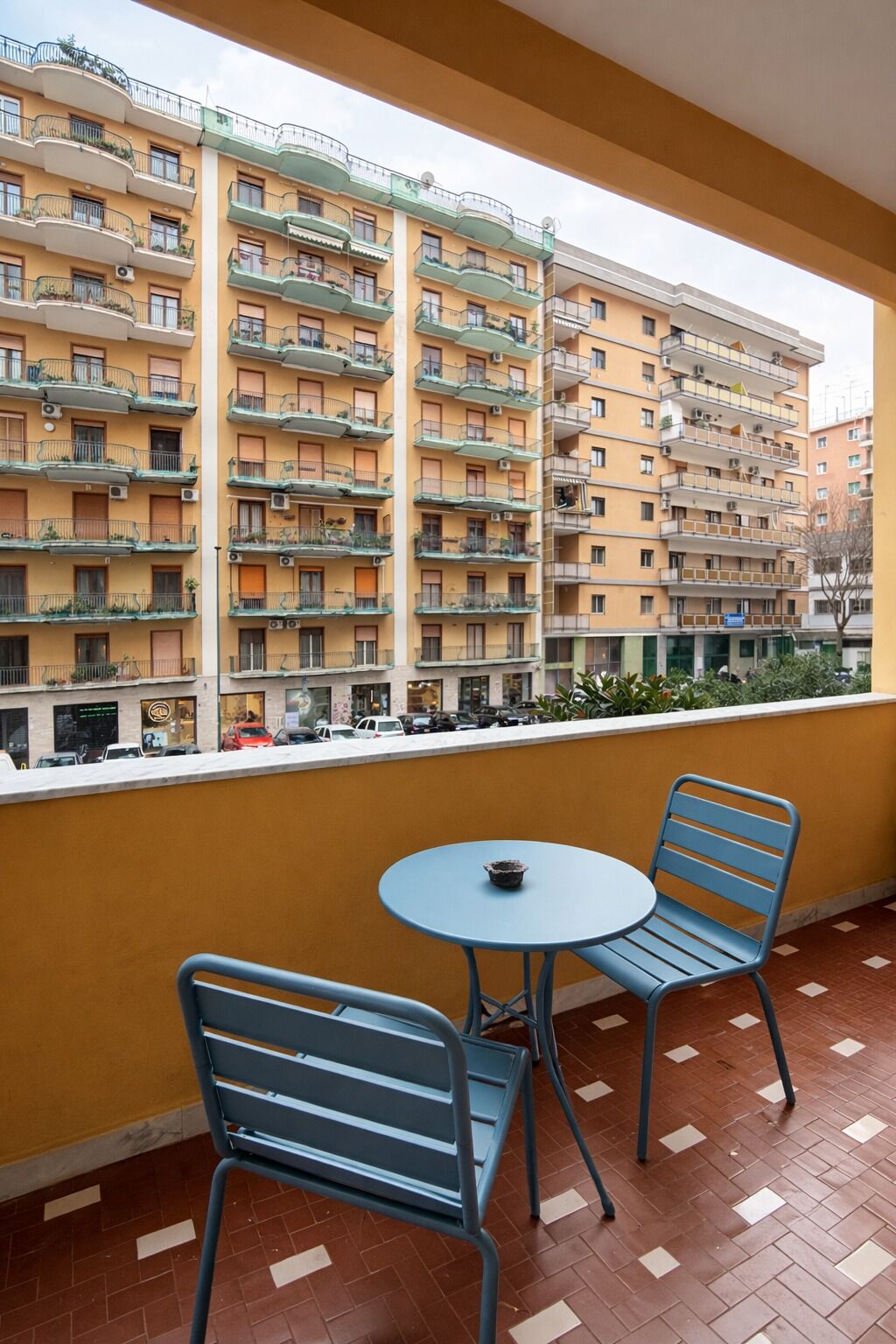 Ein Balkon mit zwei blauen Metallstühlen und einem kleinen runden Tisch, mit Blick auf ein mehrgeschossiges Gebäude mit Balkonen.
