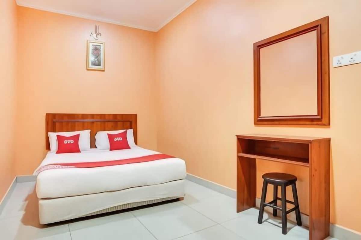 A bedroom with a bed, a wooden headboard, a red and white bedspread, a small stool, a wooden shelf, and a framed picture on the wall.