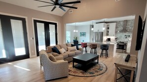A spacious living room with a ceiling fan, a patterned armchair, a sofa, and a coffee table. A kitchen area is visible in the background with modern appliances.