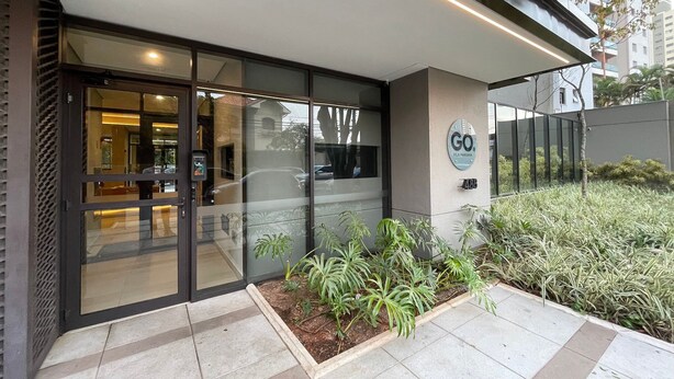 Une entrée moderne avec des portes en verre, une enseigne indiquant « GO VILA MARINA » et un petit espace jardin agrémenté de plantes.