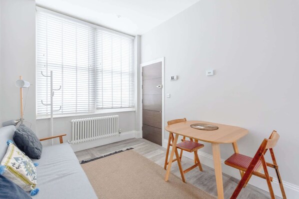 A room with a bed, a table with chairs, a radiator, and a window with blinds.