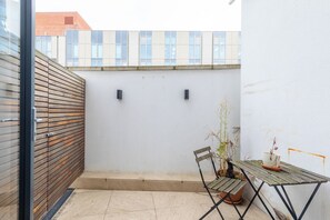Een klein terras buiten met een houten tafel en stoelen, een plant in pot en een witte muur met twee zwarte wandlampen.