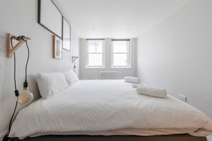 Een minimalistische slaapkamer met een groot bed, twee ramen en een radiator.