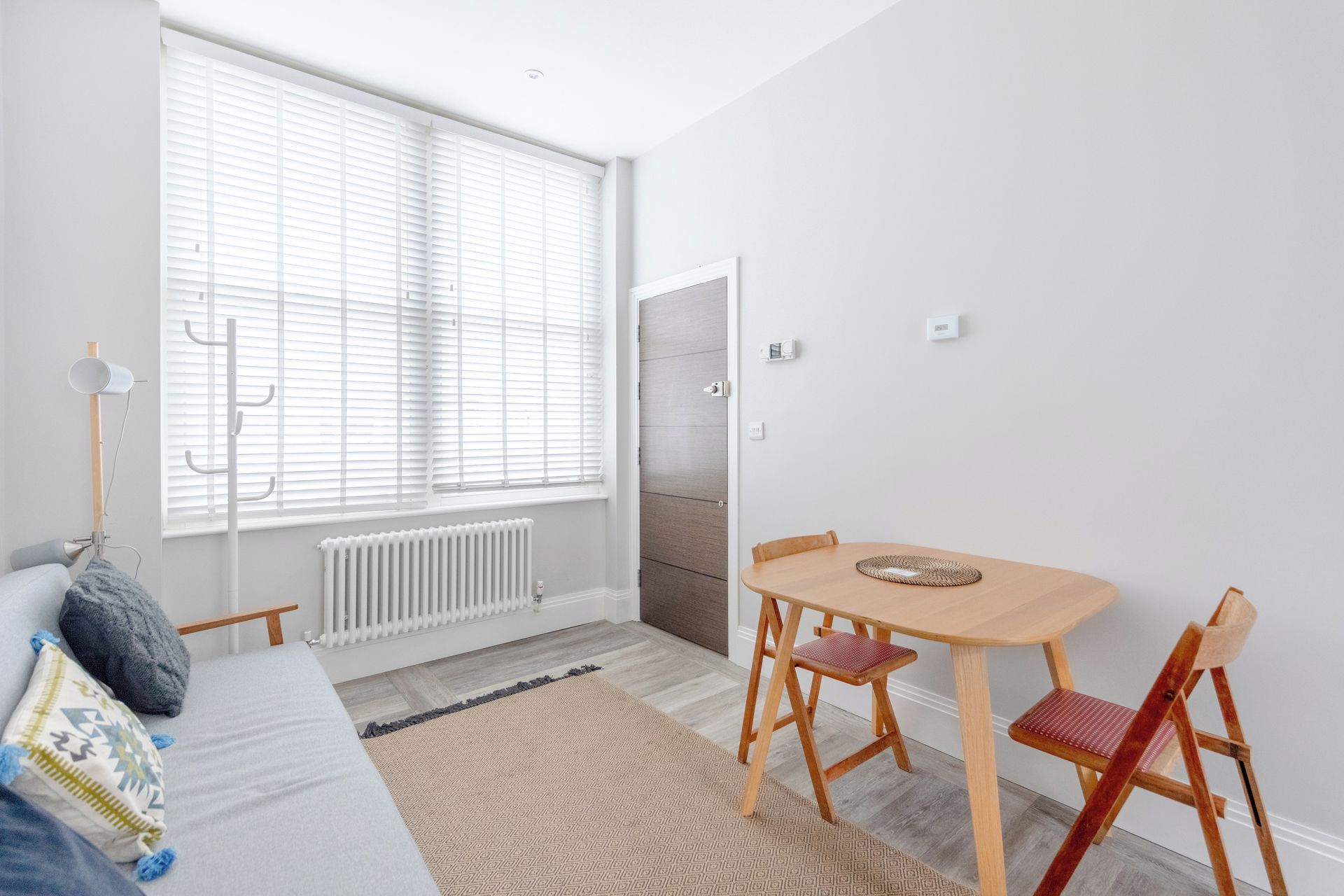 Une pièce moderne et bien éclairée, comprenant un lit, une table à manger avec des chaises, un radiateur et une fenêtre avec des stores.