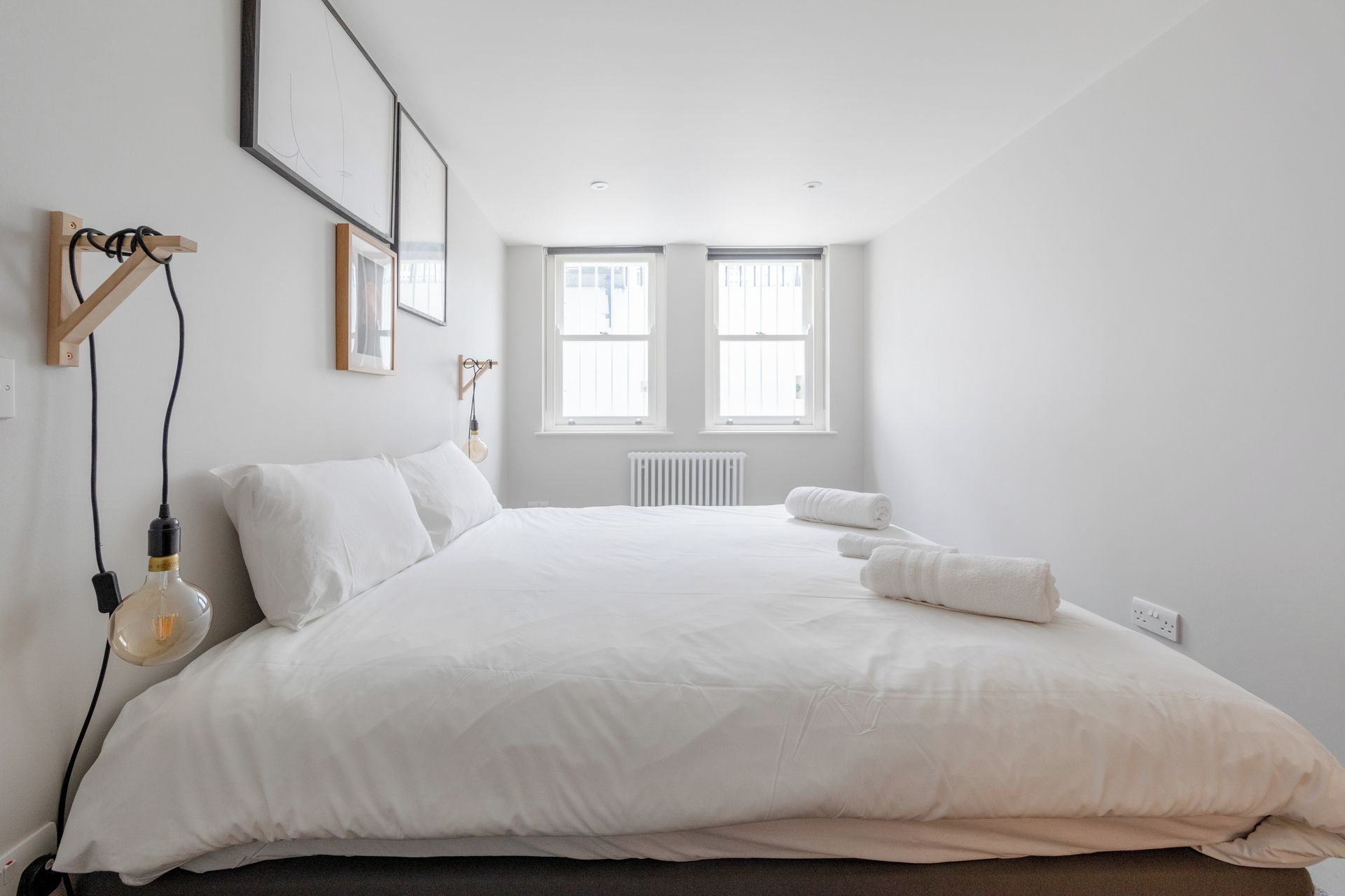 Een minimalistische slaapkamer met een groot bed, twee ramen en een radiator.