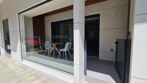 Un bâtiment moderne avec une entrée en verre, un petit espace extérieur aménagé avec deux chaises et une table, et une porte bien visible donnant accès à l’intérieur.