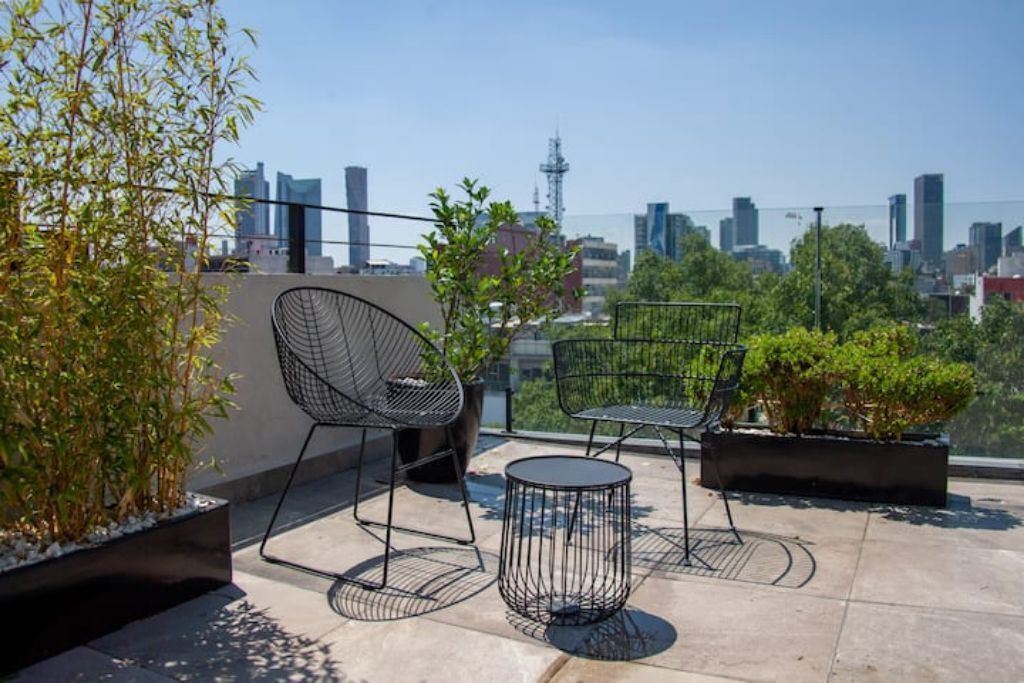 Una terraza en la azotea con dos sillas de estructura metálica negra y una pequeña mesa redonda, rodeada de plantas en macetas y vistas a un paisaje urbano.