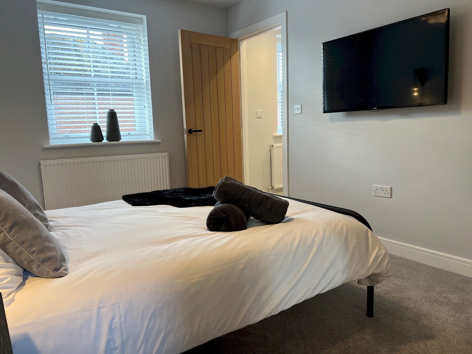 A bedroom with a bed, a television mounted on the wall, a window with blinds, and a door.