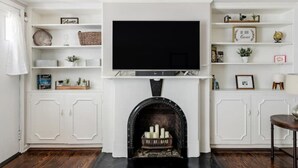A living room with a fireplace, a flat-screen TV mounted above it, and white shelving units with various decorations and books. - Pet-Friendly 1855 Garden Retreat • Savannah (Savannah)