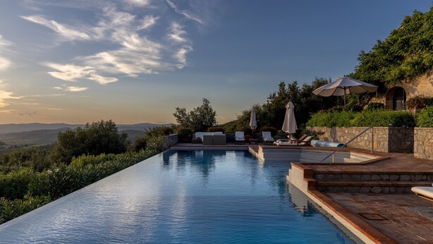 Ein Infinity-Pool mit Liegestühlen und Sonnenschirmen, umgeben von viel Grün und einer Steinmauer, mit Blick auf eine reizvolle Landschaft.