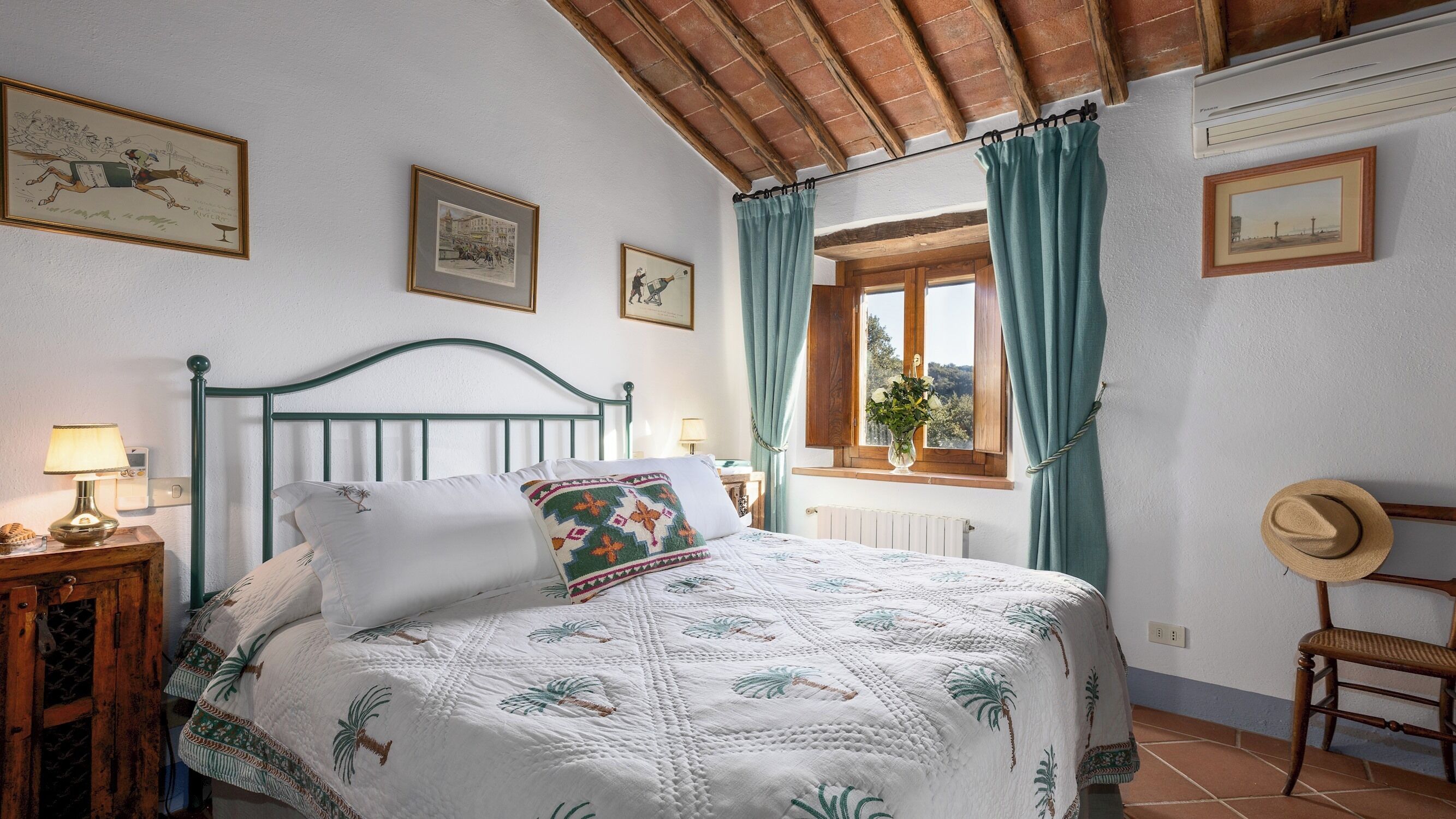 Un dormitorio con cama, mesita de noche, ventana con cortinas, una silla y cuadros en la pared.