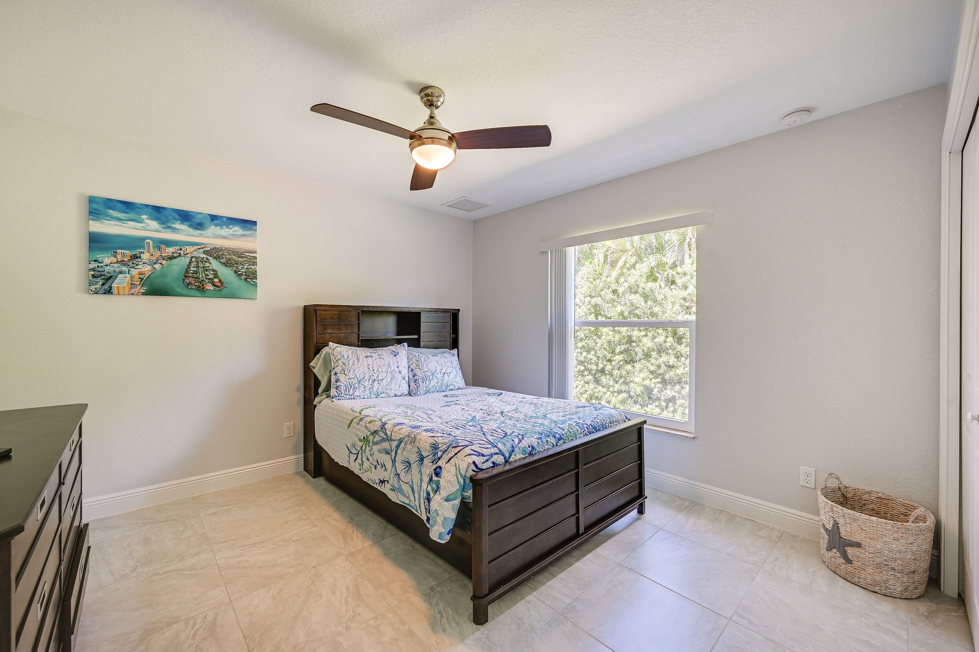 A bedroom with a bed, a ceiling fan, a window, a painting, and a small basket.