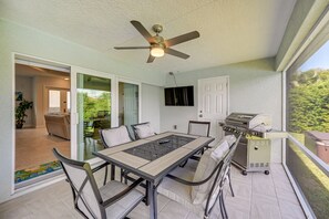 Une terrasse couverte avec une table et des chaises, un ventilateur de plafond, un barbecue et un téléviseur fixé au mur.