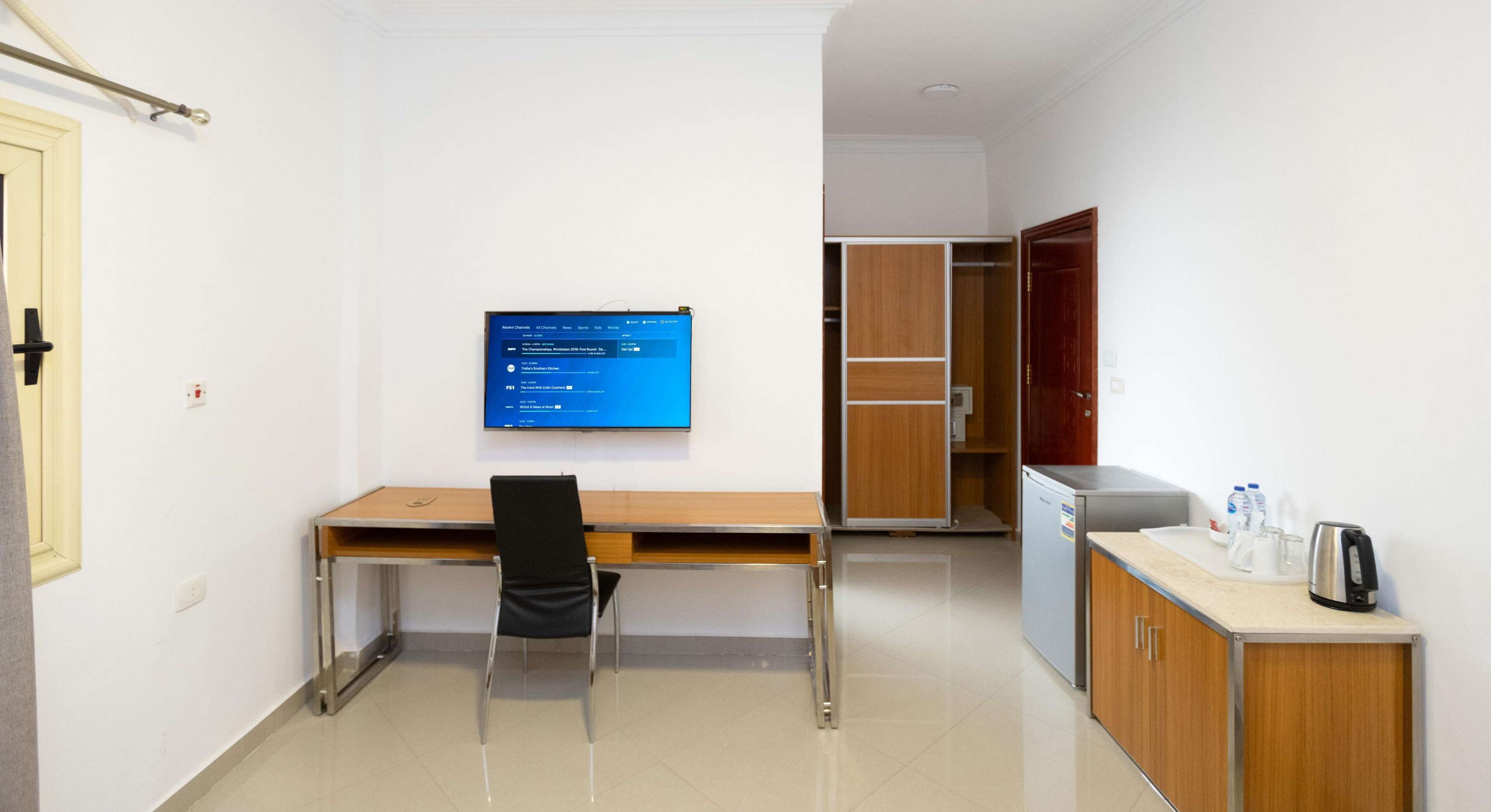 A room with a desk, chair, and a television mounted on the wall. There is a wardrobe and a kitchenette area with a refrigerator and a kettle.