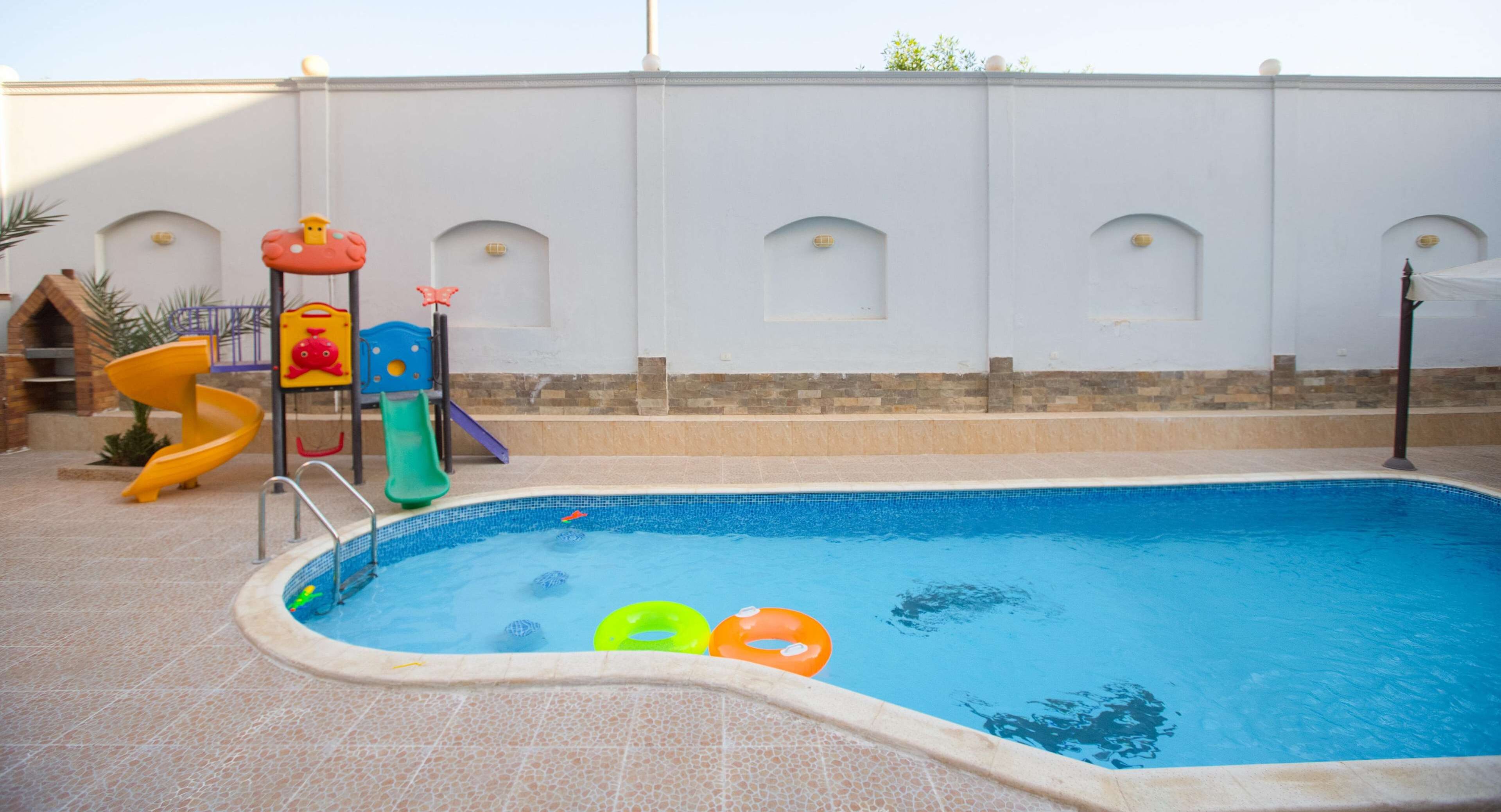 A swimming pool with a slide and play structure, surrounded by a white wall with arched windows.