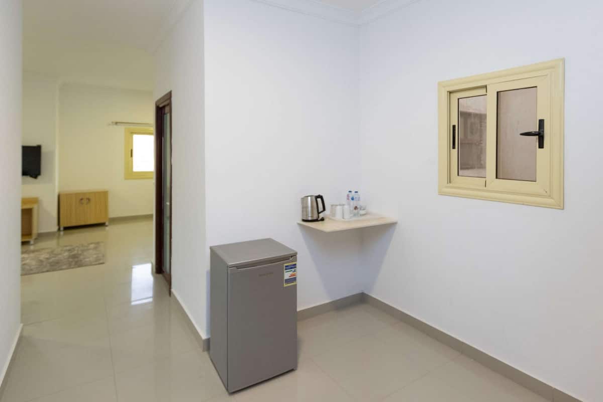A narrow hallway with a refrigerator, a small shelf with a kettle and water bottles, and a door leading to another room.