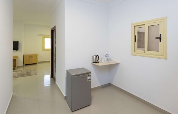 A narrow hallway with a refrigerator, a small shelf with a kettle and water bottles, and a door leading to another room. - Almanzel Sheikh Zayed  (Sheikh Zayed City)