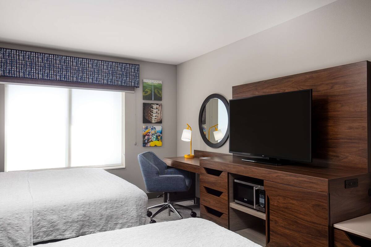 A hotel room with two beds, a desk with a TV, a lamp, and artwork on the wall.