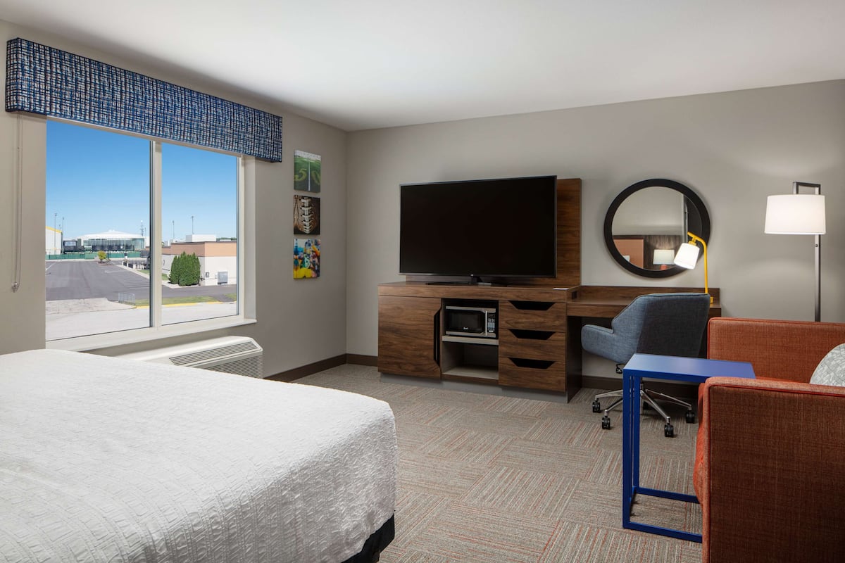 A hotel room with a large bed, a desk with a flat-screen TV, a chair, a round mirror, and a lamp.