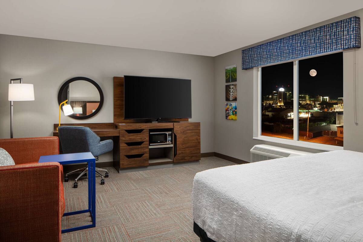A hotel room with a bed, a desk with a flat-screen TV, a chair, a desk lamp, and a large window with a city view.