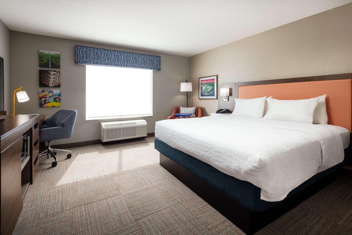 A hotel room with a large bed, a desk, a chair, a lamp, and artwork on the walls.