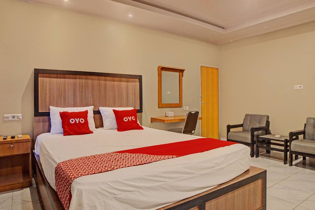 A hotel room with a large bed, two red pillows, a wooden headboard, a bedside table, a desk, a chair, and a small table.