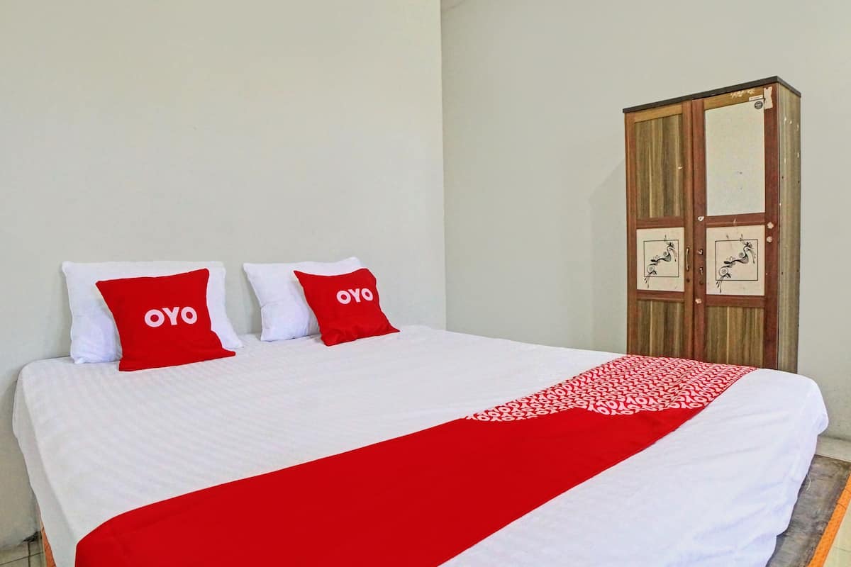 A bed with white bedding and two red pillows, a red and white patterned throw blanket, and a wooden wardrobe with a mirror and two doors.