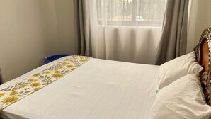 A neatly made bed with a floral bedspread, a wooden headboard, and a window with curtains. - Aval Homes-Modern 1 bedroom apartment in Bamburi (Mombasa)
