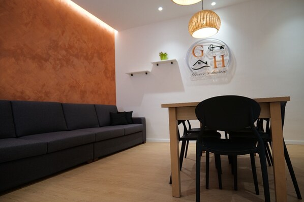 A modern living room with a dark grey sofa, a wooden dining table with a black chair, and a wall-mounted circular sign with the letters "G H" and the words "Grace's home". - B&B Affittacamere Grace's Home  (Canicatti)