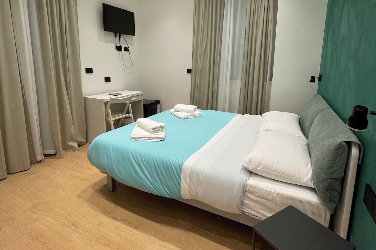 A modern hotel room with a bed, a desk, a TV mounted on the wall, and a window with curtains.