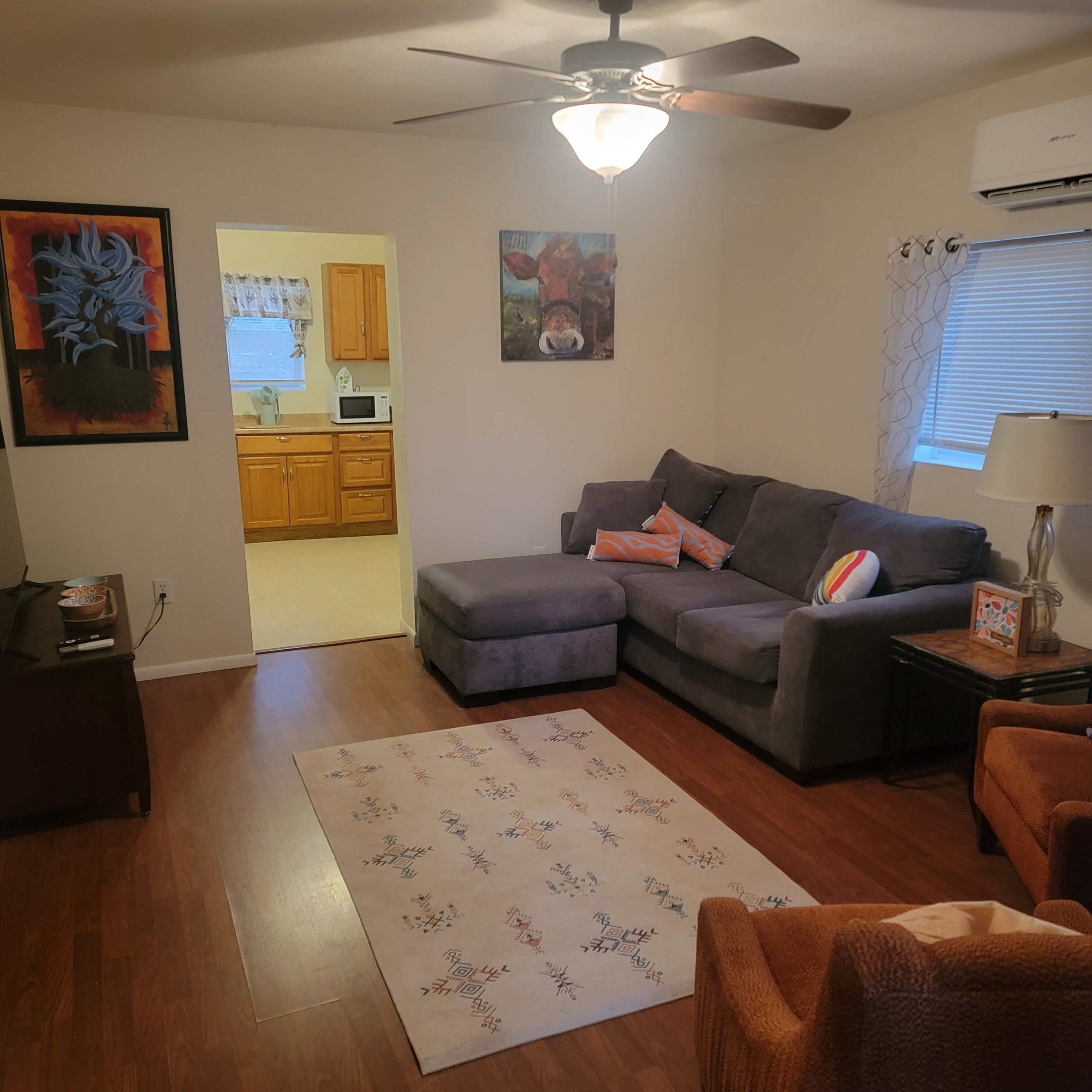 A living room with a gray sectional sofa, a small rug with Asian characters, a ceiling fan, and a kitchen in the background.