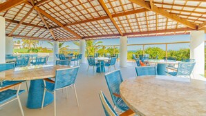 Un área de comedor al aire libre con mesas redondas y sillas azules, bajo una estructura de techo de madera.